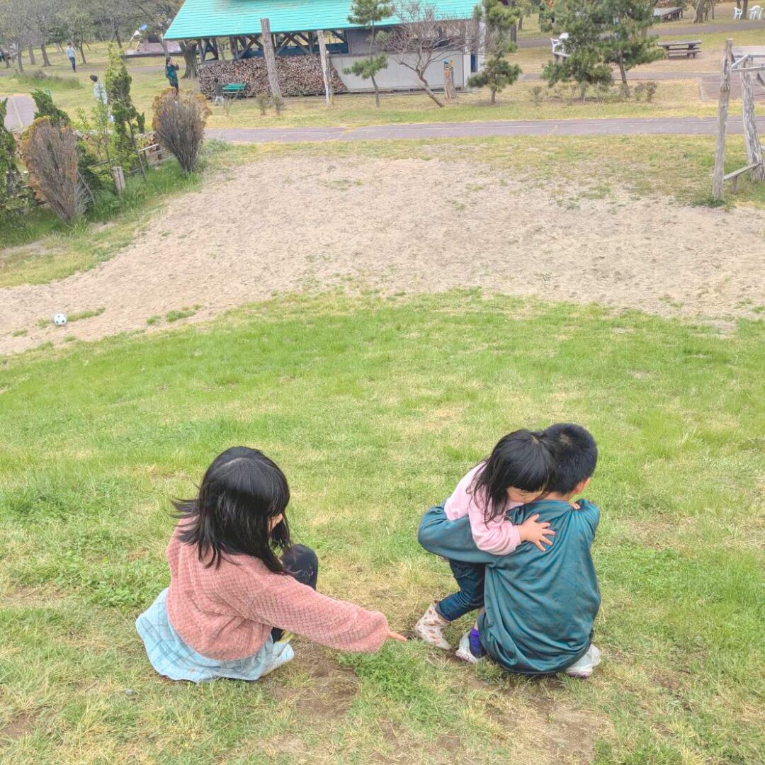 丘の上でお兄ちゃんに抱っこされている３歳の女の子と隣に座っている２年生の女の子