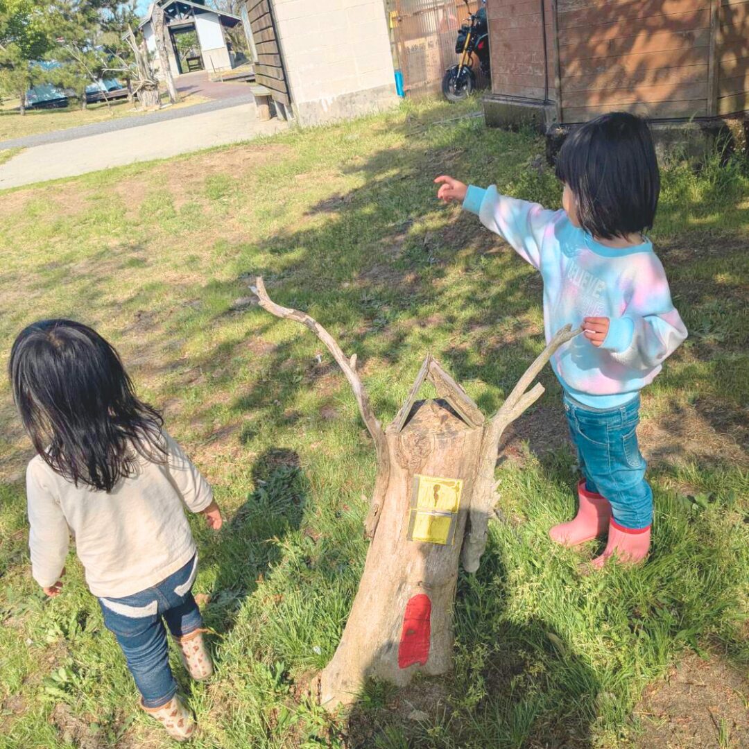 切り株にカラフルな扉が書いてあるオブジェと遊んでいる２人の子どもたちの様子