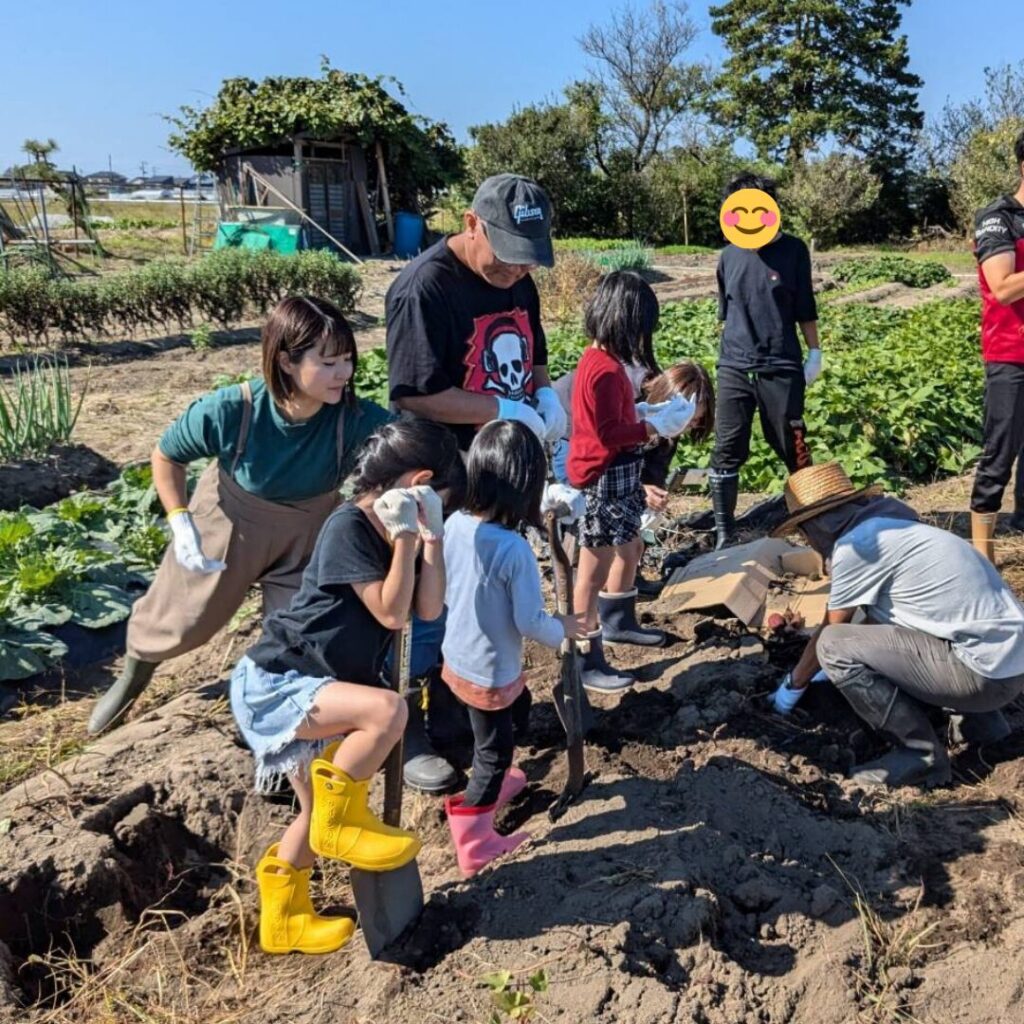 スコップを持て畑に立つこどもや大人たちが、おじさんから芋の掘り方を教えてもらっている写真